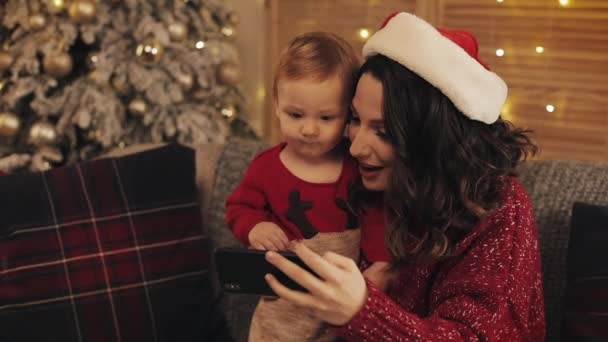 Ung mamma bär tomte hatt Sitter på soffan med sin leende lilla pojke ser glad nära julgran på varm och mysig hematmosfär Ser till hennes smartphone — Stockvideo