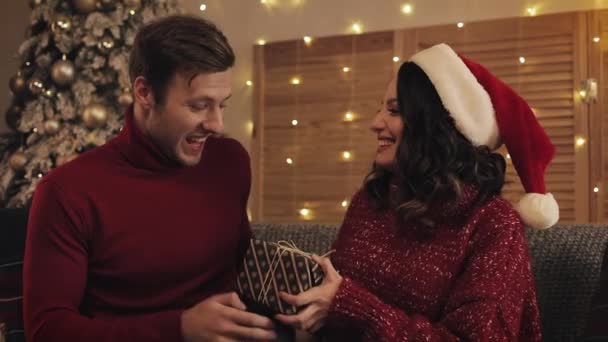 Närbild av vackra par kyssas och fru Ge hennes make närvarande Box Sitter på soffan nära julgran med skinande ljus på bakgrunden. — Stockvideo