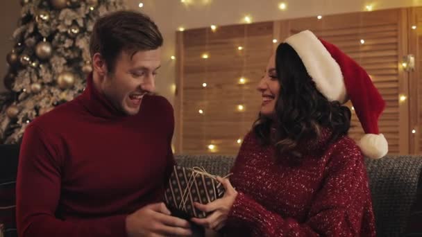 Close Up of Lovely Couple Kissing and Wife Giving her Husband Present Box Sentada en el sofá cerca del árbol de Navidad con luces brillantes al fondo . — Vídeos de Stock