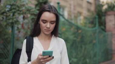 Omzunda çantasıyla, modern akıllı telefonunu kullanarak ekranı kaydıran güzel esmer kız. Kadın dışarı bakarken yana bakıyor ve gülümsüyor. Yavaş Hareket.