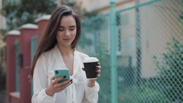 Hübsche junge lächelnde Brünette, die auf der gemütlichen Altstadtstraße spaziert, während sie ihr Handy benutzt und Kaffee aus einer Papptasse trinkt und auf den Bildschirm schaut. Kommunikations- und Technologiekonzept. — Stockvideo