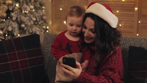 Junge schöne Mutter mit Weihnachtsmütze auf dem Sofa sitzend mit ihrem niedlichen kleinen Jungen, der glücklich lächelnd in der Nähe des Weihnachtsbaums in warmer und gemütlicher Atmosphäre zu Hause einen Videoanruf macht — Stockvideo