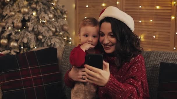 Schöne junge lächelnde Mama mit ihrem süßen kleinen Jungen, der auf der Couch im Wohnzimmer in der Nähe des Weihnachtsbaums sitzt und Selfie oder Videobotschaft von Urlaub und Neujahr macht. — Stockvideo