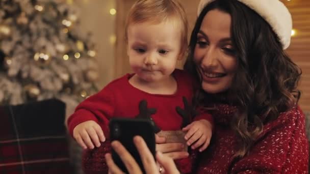 Lächelnd schöne Mutter mit ihrem kleinen Sohn bei gemütlichen Weihnachten zu Hause Hintergrund sitzt auf dem Sofa mit Videoanruf chatten Konzept von Urlaub und Neujahr aus nächster Nähe. — Stockvideo