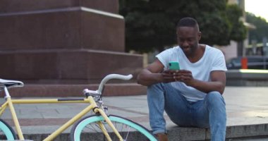 İyi görünümlü Afro-Amerikalı genç adam akıllı telefonunu kullanıyor, mesajlar yazıyor, heyecanlı görünüyor şehrin arka planında Stylish Bisikleti 'nin yanında otururken. Ön Görünüm.
