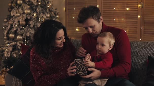 Joven familia sonriente con bebé niño mirando feliz se sienta en el sofá cerca del árbol de Navidad en la cálida y acogedora atmósfera del hogar sosteniendo una caja de regalo. Concepto de Vacaciones y Año Nuevo . — Vídeos de Stock