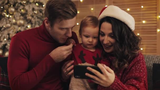 Jovem Família Alegre de Mãe Pai e Menino Senta-se no Sofá perto da Árvore de Natal com Luzes Brilhantes no Fundo Sorrindo Fazendo Vídeo Mensagem ou Vídeo Chamada — Vídeo de Stock