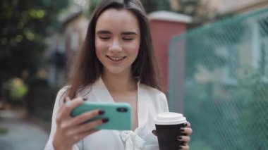 Kahverengi Saçlı Çekici Genç Kız Kâğıt Kupa 'dan Kahve İçiyor ve Akıllı Telefonunu Yatay Tutarak Video İzliyor. Rahat şehir sokağında yürüyen kadın..