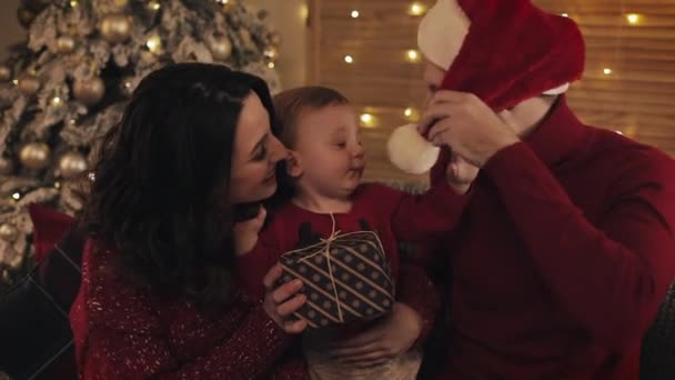 Glückliche kaukasische Mutter Vater und kleiner Sohn in Winterpullovern sitzen im gemütlichen Wohnzimmer auf der Couch in der Nähe des Weihnachtsbaums lächelnd und haben Spaß dabei, eine Geschenkschachtel in der Hand zu halten — Stockvideo