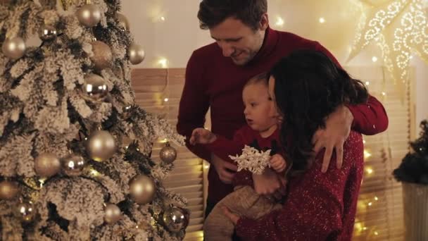 Feliz familia sonriente con el pequeño bebé de pie cerca del árbol de Navidad en la acogedora sala de estar, mamá sostiene y abraza al bebé, el esposo abraza y le da a su esposa un concepto de beso de vacaciones familiares y año nuevo de cerca . — Vídeo de stock