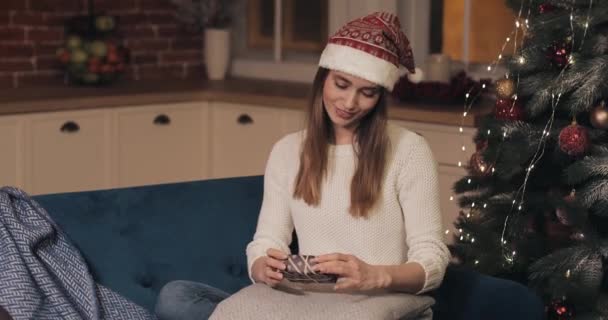 Jóvenes mujeres hermosas con sombrero de santas sentadas en un sofá en el acogedor fondo de Navidad sosteniendo una pequeña caja de regalos. Concepto de vacaciones y año nuevo de cerca  . — Vídeo de stock
