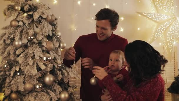 Jonge vriendelijke glimlachende familie van mama papa en hun baby zoon versieren kerstboom in hun woonkamer op zoek gelukkig. Begrip gezinsvakantie en nieuwjaar. — Stockvideo