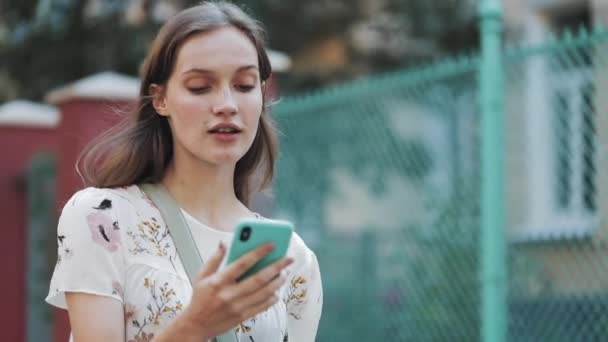 Primer plano de la hermosa mujer joven con vestido blanco de la flor con el bolso cruza su hombro usando su teléfono inteligente. Chica mirando sorprendido y diciendo que sí caminando cerca de la cerca de la turquesa sonriendo . — Vídeo de stock