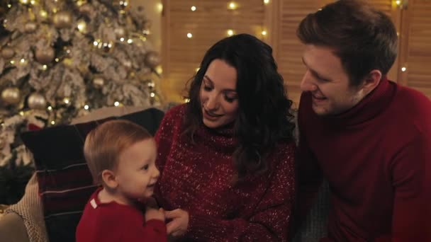 Primer plano de la feliz familia sonriente sentada en el sofá cerca del árbol de Navidad, padres jugando con su adorable hijo bebé, papá abraza a mamá. Concepto de vacaciones en familia y Año Nuevo . — Vídeos de Stock