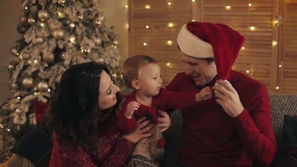 Happy Young Parents and their Cute Todler Boy Sentado en las rodillas de los papás cerca de Christmas Tree Smiling. Papá juega con el bebé usando su sombrero en el ambiente cálido y acogedor del hogar . — Vídeos de Stock