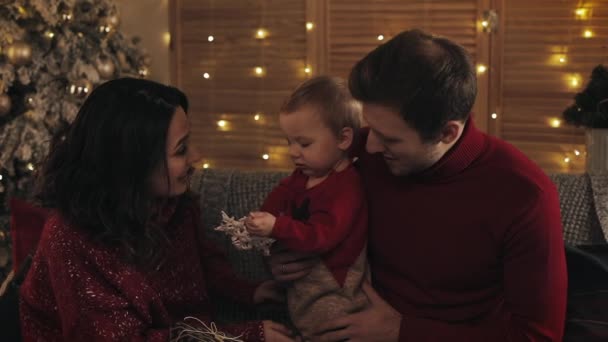 Jeune famille avec bébé garçon regardant heureux s'assoit sur le canapé près de l'arbre de Noël à l'atmosphère chaleureuse et confortable de la maison, enfant tient jouet flocon de neige. Concept de vacances et Nouvel An . — Video