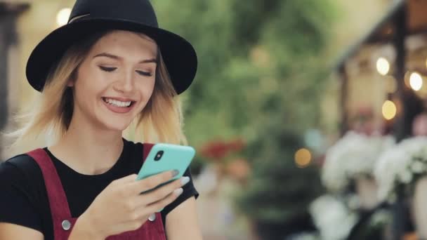 Retrato de una jovencita sonriente bastante rubia usando un sombrero negro usando su teléfono inteligente de pie en la calle City . — Vídeos de Stock