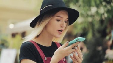 Siyah Şapkalı Güzel Sarışın Kız Akıllı Telefon Ses Komutanını kullanarak Şehrin Sol Tarafının Görünüşünde Yürüyor.