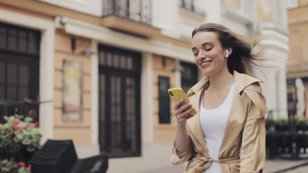 Jovem sorrindo muito caucasiano menina vestindo fones de ouvido andando na rua segurando telefone em sua mão e digitando tempo ventoso . — Vídeo de Stock