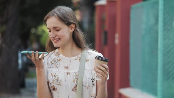 Menina bonita nova vestindo vestido de flor branca com saco Cruz seu ombro segurando xícara de café e seu Smartphone e fazendo mensagem de voz ou usando o comandante de voz. Mulher caminhando na rua . — Vídeo de Stock