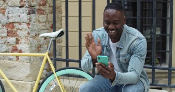 Веселый афроамериканец с видеозвонком через смартфон. Парень машет и улыбается, сидя возле стильного велосипеда со старым металлическим фэнсом на заднем плане . — стоковое видео