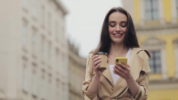 Ελκυστική νεαρή κοπέλα Χρησιμοποιώντας Smartphone και χαμογελώντας κρατώντας ένα φλιτζάνι χαρτί πίνοντας καφέ στέκεται στο παρασκήνιο της παλιάς πόλης άποψη παρακάτω. — Αρχείο Βίντεο