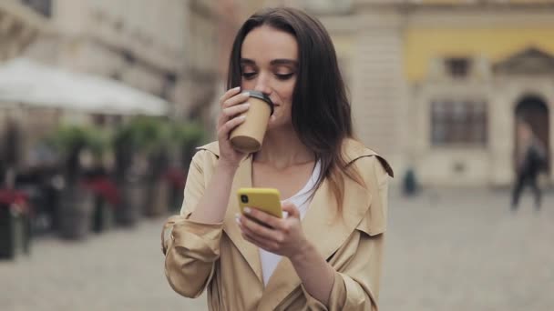Mladý atraktivní dívka pití kávy smích pomocí svého smartphonu a psaní stojící na pozadí města. — Stock video