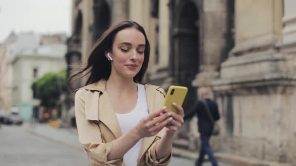 Schöne glückliche junge Mädchen trägt drahtlose Kopfhörer macht einen Videoanruf lächelnd reden zu Fuß entspannt auf der Straße Stadt Hintergrund Nahaufnahme. — Stockvideo
