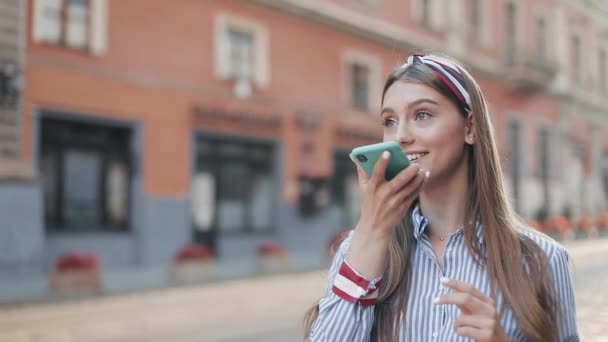 Jeune belle femme avec bandeau dessus portant une robe rayée à la mode en utilisant son Smartphone Voice Commander ou en faisant un message vocal. Fille défilant téléphone marche sur la rue de la ville . — Video