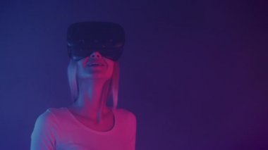 Sanal gerçeklik gözlüğü takan mutlu, gülümseyen sarışın kızın yakın portresi. Neon ışıklı renklerin olduğu odada duruyor. Gelecek vaat eden arka plan..