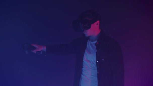 在虚拟现实眼镜中迷人的男人用控制机画出抽象线条和人物形象，站在有霓虹灯照明色彩的未来背景的房间里. — 图库视频影像