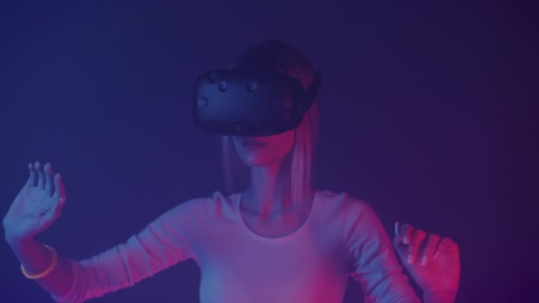 VRヘッドセットを装着した白人の少女の密室、空中で手を動かし、抽象的なネオンカラーの背景を持つ部屋に立つ。未来構想. — ストック動画
