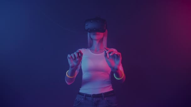 VRヘッドセットを身に着けている若いブロンドの髪の女の子,タッチVR画面,タイピング,抽象ネオン照明の色の背景と部屋に立って.未来、 VRコンセプト. — ストック動画