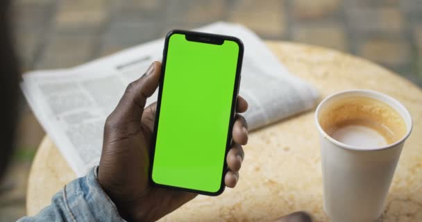 Lviv, Ukraine - Le 09 août 2019 : Des mains d'Afro-Américain assis à table avec du papier journal et une tasse de café dessus, tenant un smartphone avec écran vert et buvant du café — Video