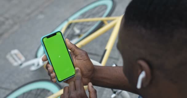 Lviv, Ucrania - 09 de agosto de 2019: Primer plano del hombre afroamericano con auriculares sosteniendo su teléfono inteligente tocando la pantalla verde y mirándola mientras está sentado en los escalones cerca de Bike at the City Background . — Vídeos de Stock