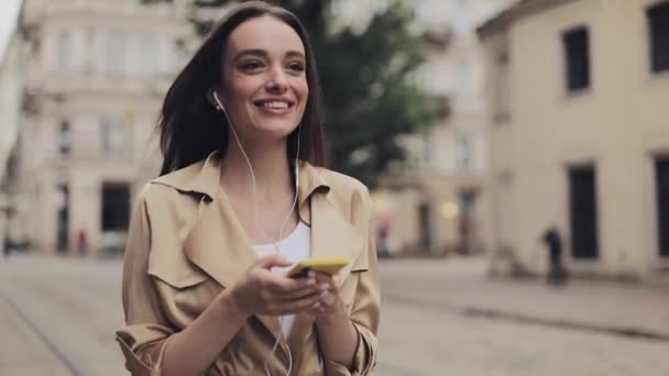 Привлекательная девушка в наушниках слушает музыку с помощью смартфона и улыбается, гуляя по городу . — стоковое видео
