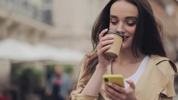 Attraktives junges Mädchen tippt auf ihr Smartphone und lächelt mit einem Pappbecher, der Kaffee trinkt, vor dem Hintergrund der Altstadt aus nächster Nähe . — Stockvideo