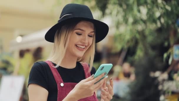 Joven rubia caucásica usando un sombrero negro usando su smartphone escuchando una voz Messege de pie en la calle de la ciudad . — Vídeos de Stock