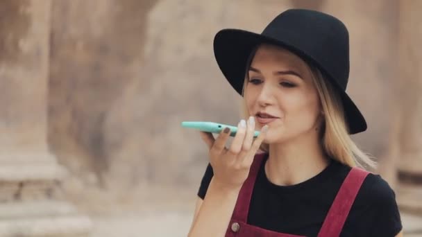 Joven chica rubia sonriente atractiva con un sombrero negro usando su reconocimiento de voz de teléfono inteligente caminando por la calle de la ciudad . — Vídeos de Stock