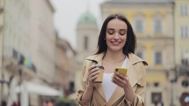 Jong mooi meisje drinken koffie glimlachen met behulp van haar smartphone en typen wandelen op City Street. — Stockvideo