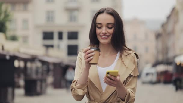 Söt ung kvinna bär Beige Trench Använda sin smartphone och ler håller en pappersmugg dricka kaffe promenader på Gamla stan bakgrund. — Stockvideo