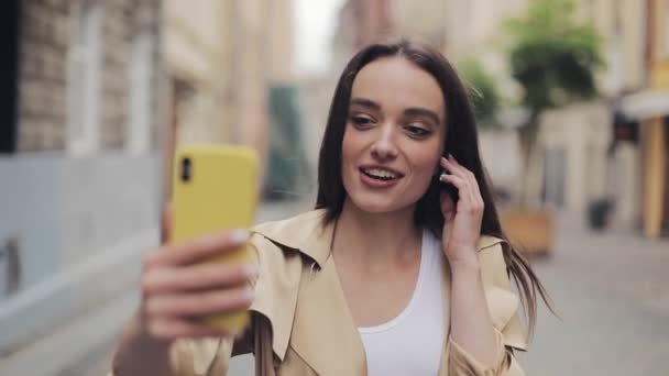 Hermosa joven sonriente usando auriculares tocándolos haciendo una videollamada sosteniendo el teléfono hablando verticalmente y de pie en el fondo de la calle de la ciudad . — Vídeo de stock