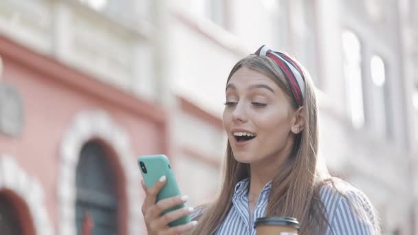 Söt kvinna klädd i blå och vit randig klänning med hjälp av app på smartphone gå på den gamla stadsgatan. Vacker flicka som har goda nyheter på smartphone. — Stockvideo