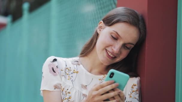 Close Up di giovane sorridente ragazza affascinante con i capelli castani e occhi blu con lentiggini in piedi appoggiata a Fance utilizzando il suo smartphone chat digitando cercando sorpreso dicendo Wow . — Video Stock
