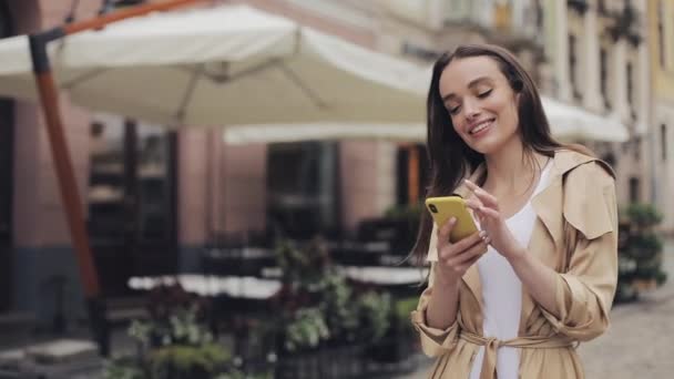 Atraktivní mladá dívka nosí sluchátka poslech hudby pomocí svého chytrého telefonu a úsměv stojící na pozadí města. — Stock video