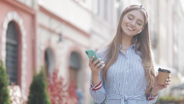 Junge lächelnde Frau mit braunen Haaren in stylischem gestreiftem Hemd und Stirnband, die zufrieden und glücklich mit ihrem Smartphone und Kaffeetasse in der Hand aussah. Mädchen zu Fuß auf der alten Stadtstraße. — Stockvideo