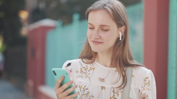 Close-up portret van jong aantrekkelijk meisje met bruin haar dragen oortelefoons genietend luisteren naar muziek glimlachen sluit ogen staande op straat. — Stockvideo