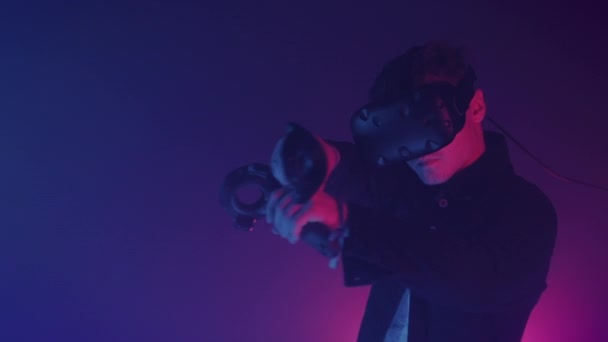 Молодой человек носит виртуальную реальность Heaadset держа джойстики контроллеры указывая, что пистолет. Игра съемки игры в комнате с неоновой освещения Цвета фона — стоковое видео