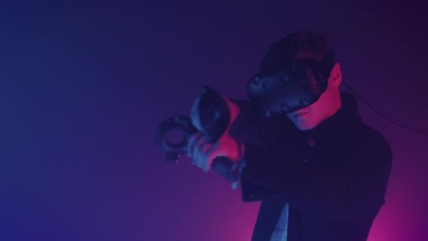 Jovem Vestindo Realidade Virtual Heaadset Segurando Joysticks Controladores Apontando essa arma. Jogar jogo de tiro na sala com Neon Iluminação Cores Fundo — Vídeo de Stock