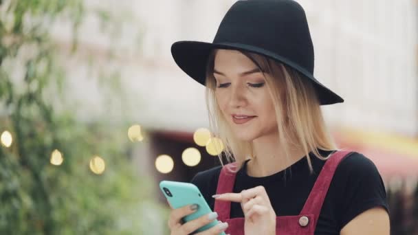 Молода дівчина, яка посміхається і усміхається, одягається в чорну капелюшку на своєму смартфоні і стоїть на задньому плані міста. — стокове відео